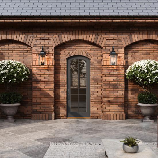 Brick courtyard with arched masonry details, black-framed entry door, decorative wall lanterns, large flowering topiary planters, and stone tile patio.