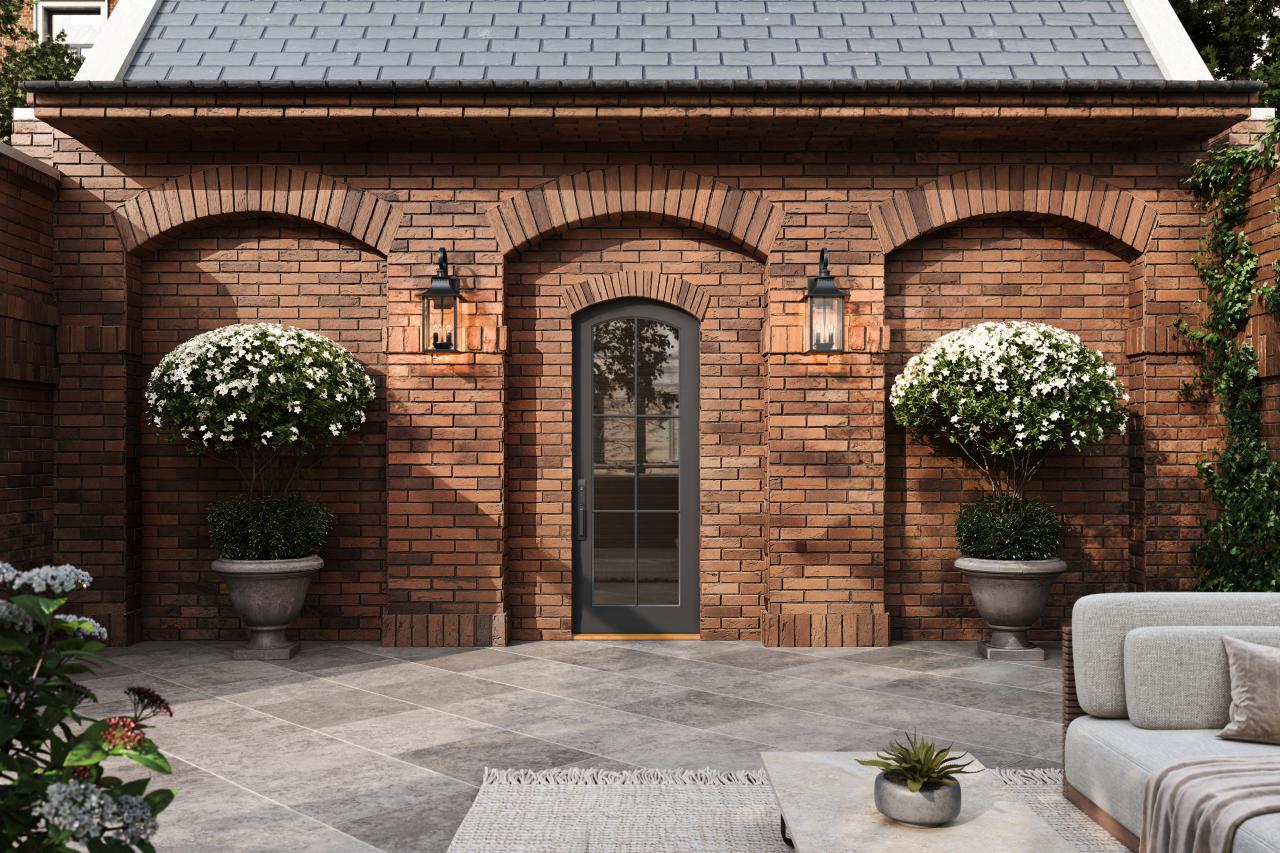 Brick courtyard with arched masonry details, black-framed entry door, decorative wall lanterns, large flowering topiary planters, and stone tile patio.