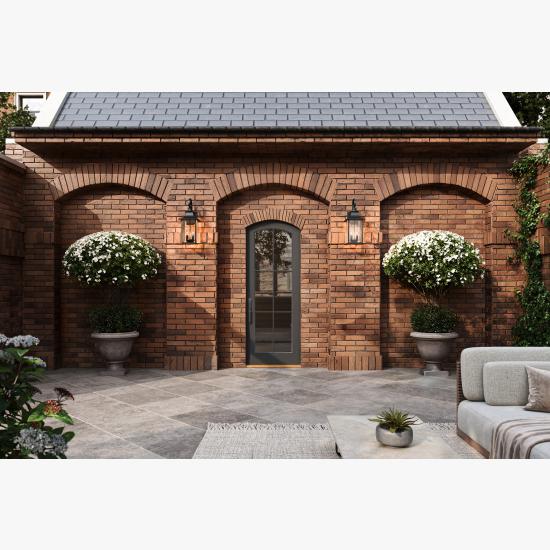 Brick courtyard with arched masonry details, black-framed entry door, decorative wall lanterns, large flowering topiary planters, and stone tile patio.